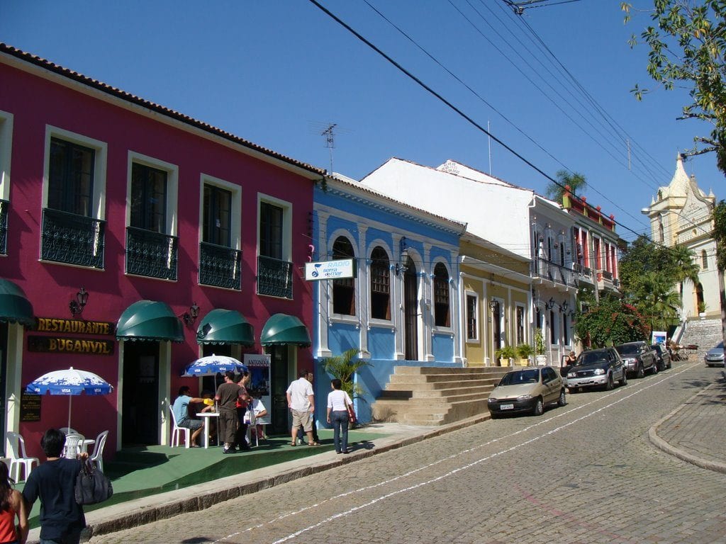 A imagem é de uma rua de antonina, com casas e comércios com paredes coloridas, alguns carros estacionados ao lado e uma igreja antiga ao fundo. 