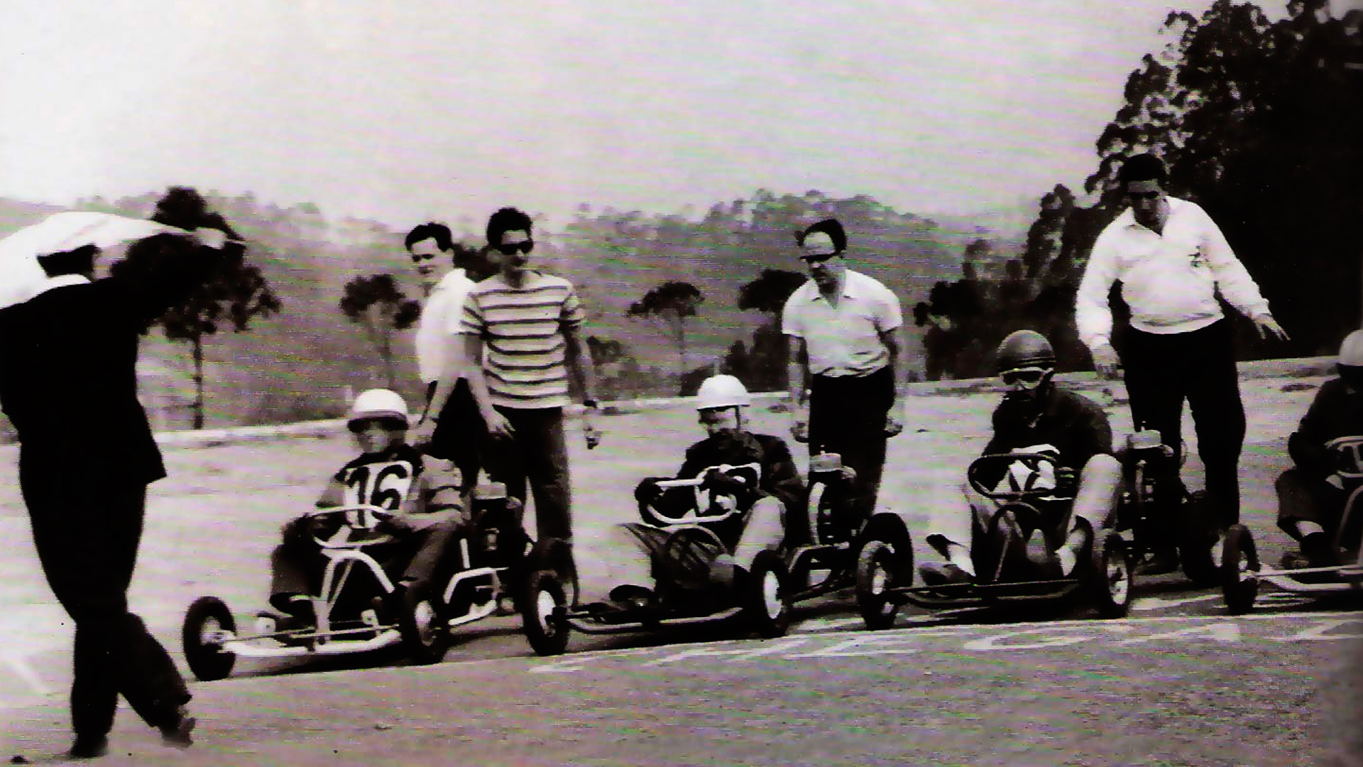 Imagem histórica de uma corrida de kart bem antiga. Na imagem tem 4 pilotos, além de 5 pessoas próximas aos karts.