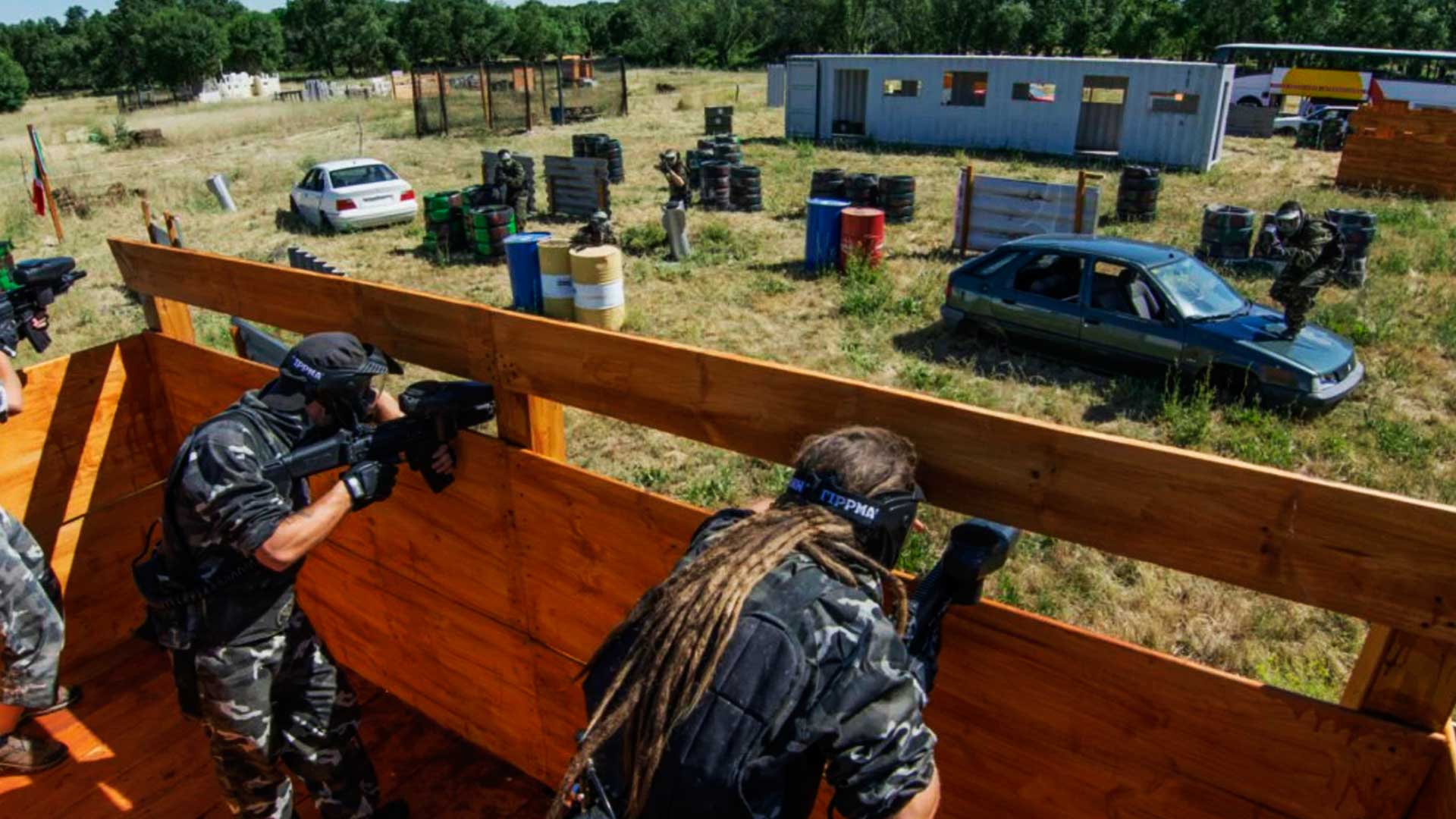 Campo de paintball outdoor, onde duas pessoas estão dentro de uma estrutura de madeira e mirando em dois adversários que estão fora. Um dos que estão fora, está em cima de um carro, já o outro está exporte, ao lado de barris.