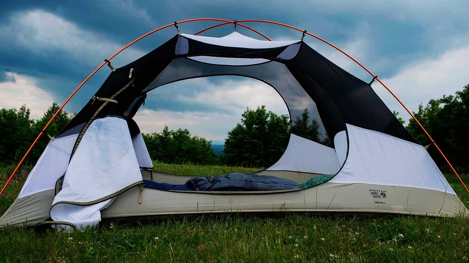 Barraca de camping montada na grama, perto de diversas árvores e com um céu bem azul e com bastante nuvens.