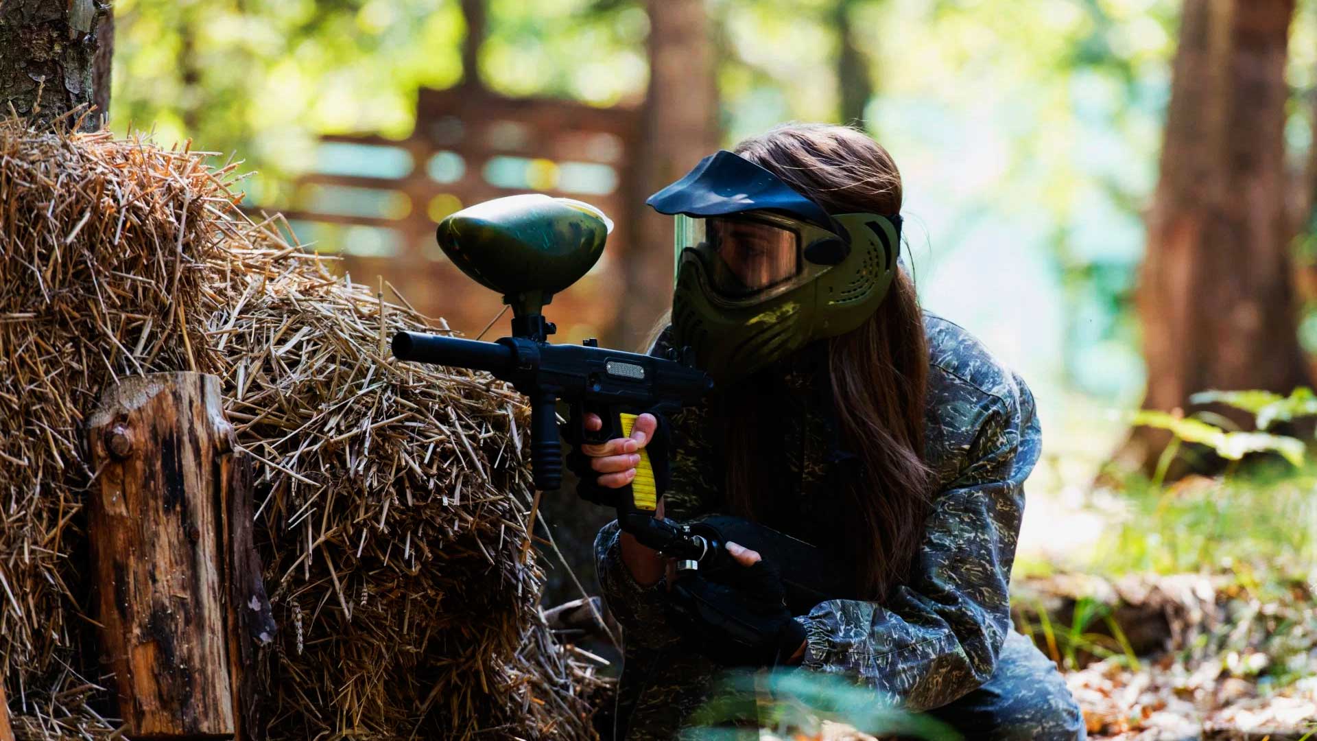 Jogadora de paintball agachada atrás de um entulho de madeiras, mirando para frente.