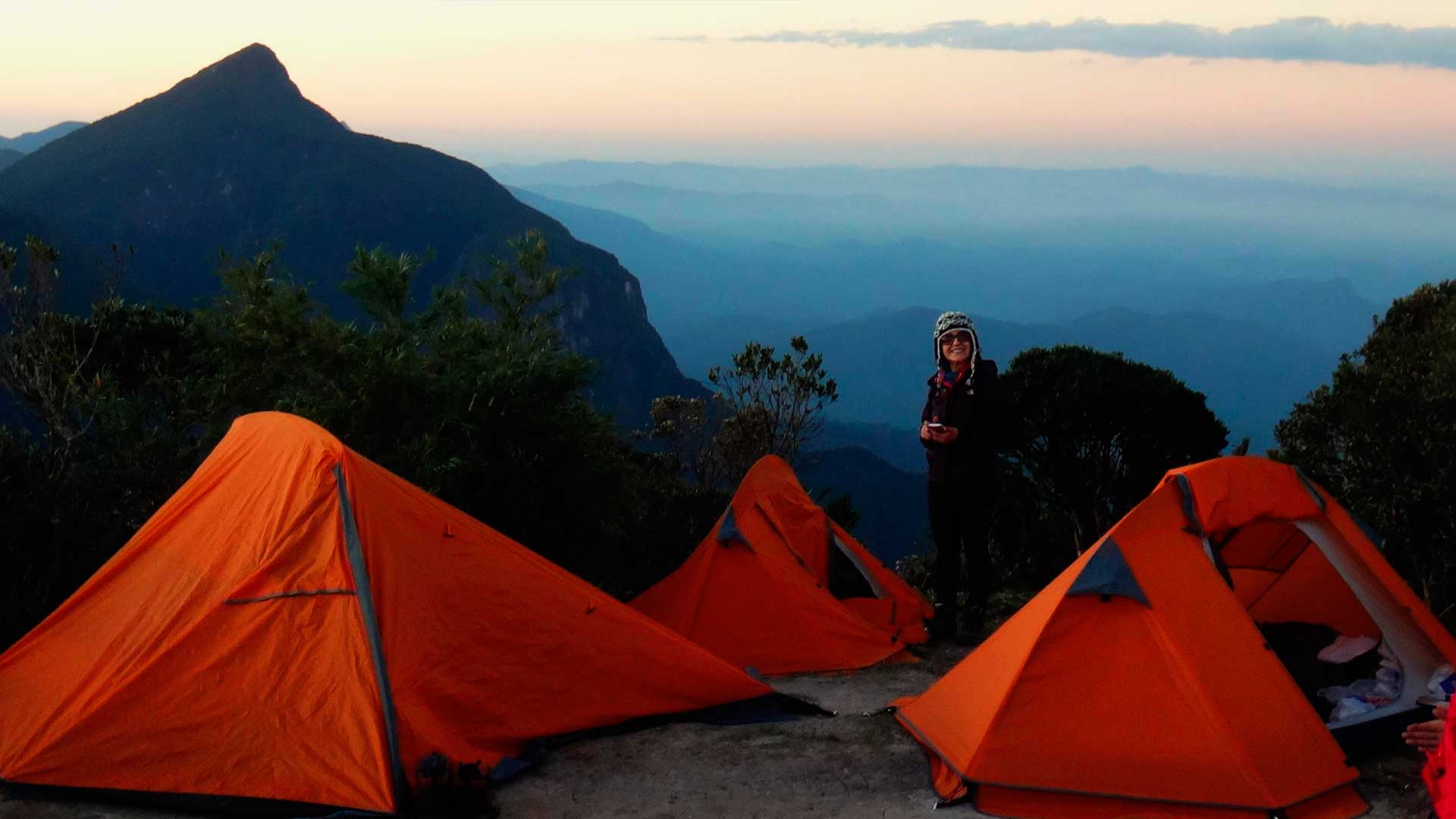 Mulher acampando no pico de uma montanha, com 3 barracas laranjas montadas. Ao fundo vemos outras montanhas e nuvens.