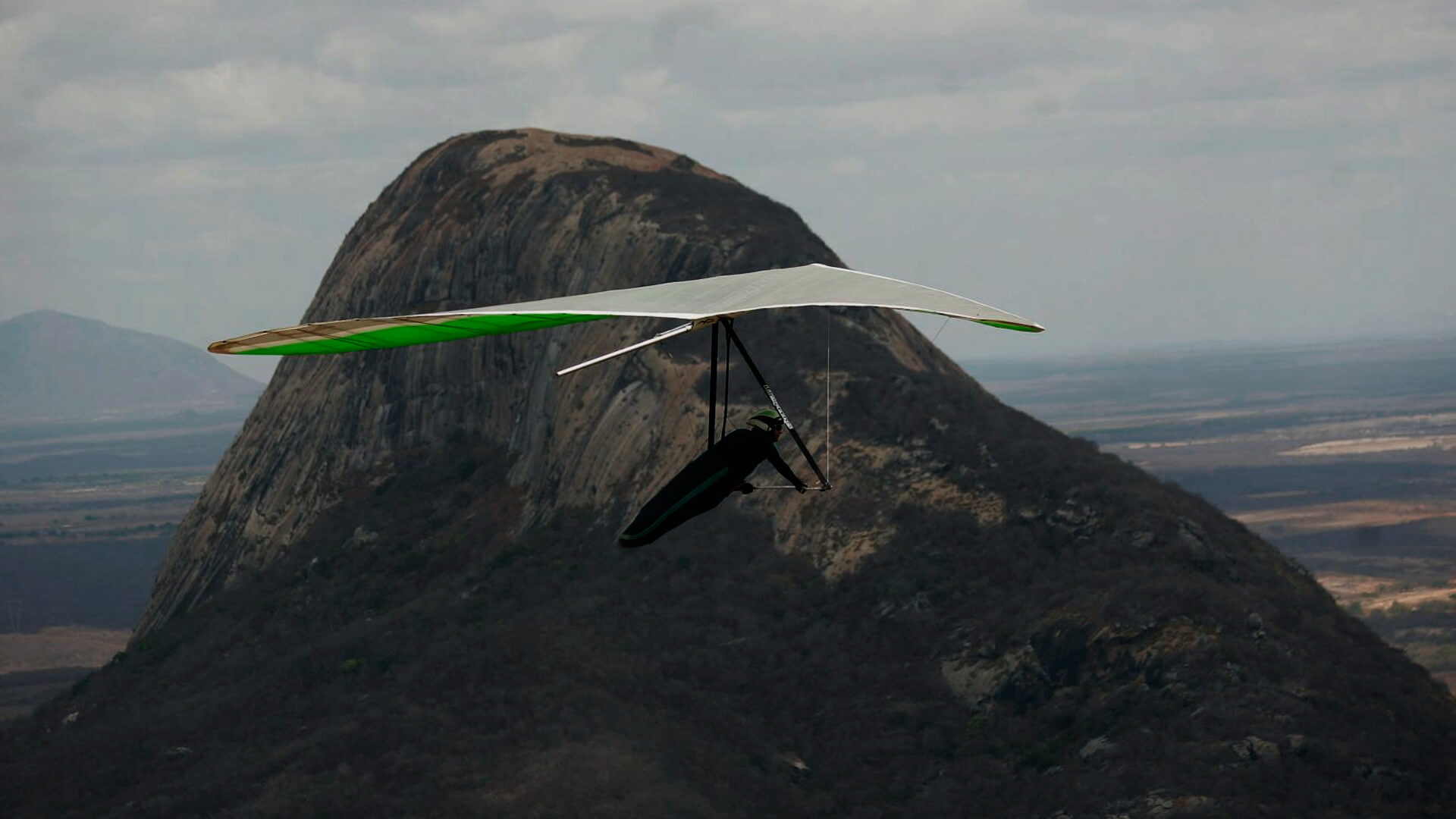 Homem praticando parapente com uma vista linda. Ao fundo uma montanha e também um campo a perder de vista. O dia está nublado, com muitas nuvens.