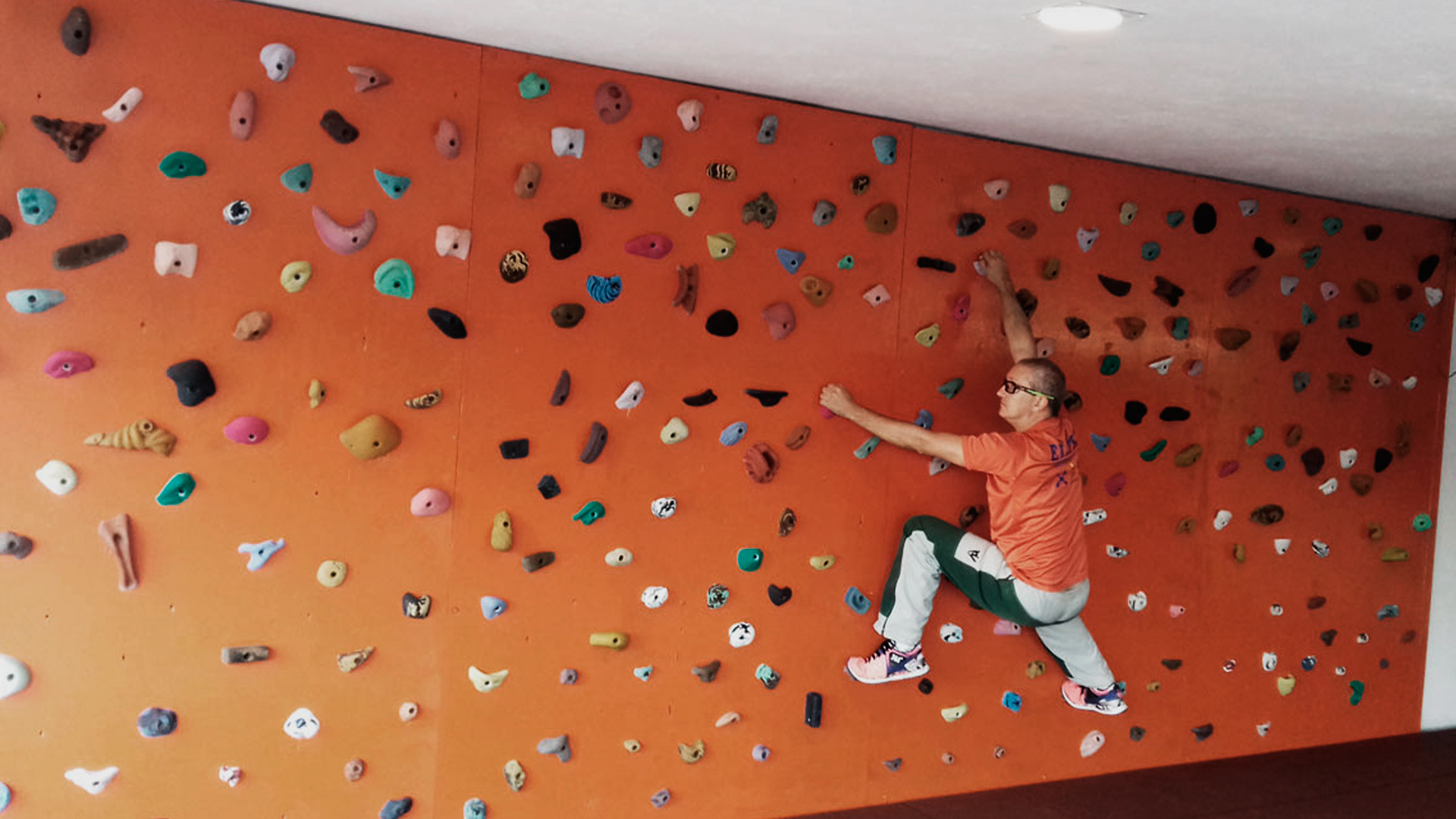 Homem praticando escalada indoor em uma parede laranja.