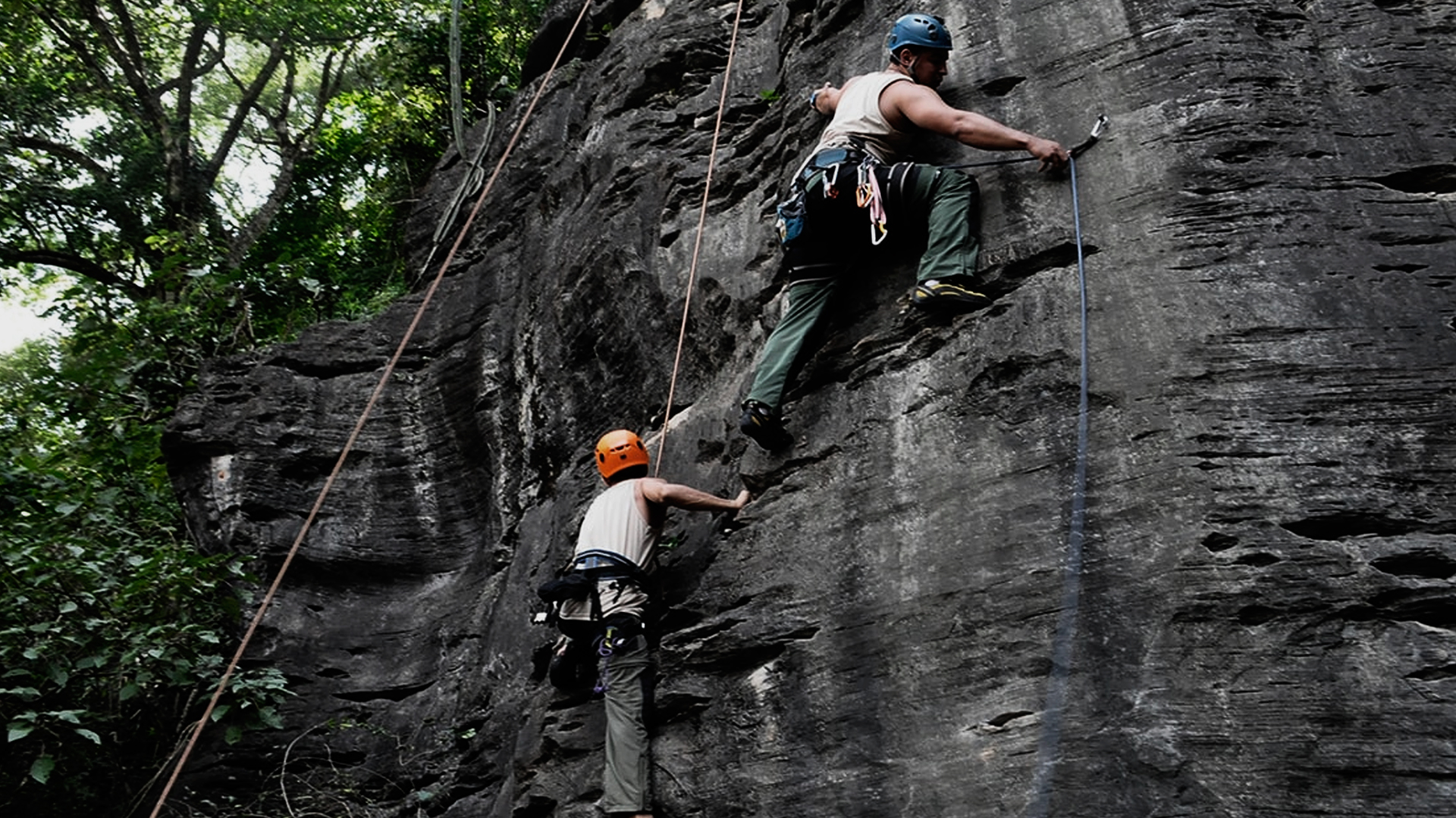 Duas pessoas praticando escalada outdoor. Ambas com capacete de segurança e também cordas de segurança.