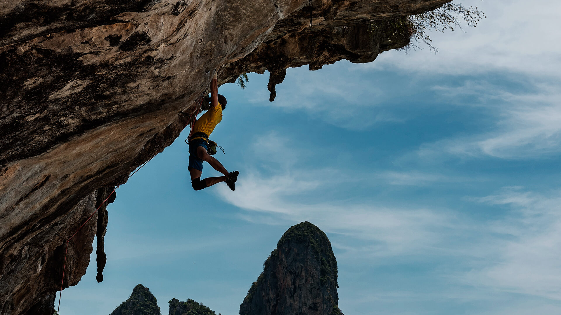 Homem praticando escalada em uma montanha. Está pendurado na montanha, no fundo um céu bem azul.