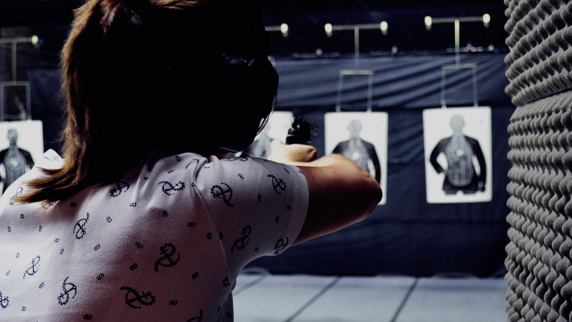 Mulher mirando em um alvo dentro de um estande de tiro. Ela esta de costas, com protetor auricular e óculos de proteção. Está mirando com uma pistola, que está apontada para o alvo do estande.