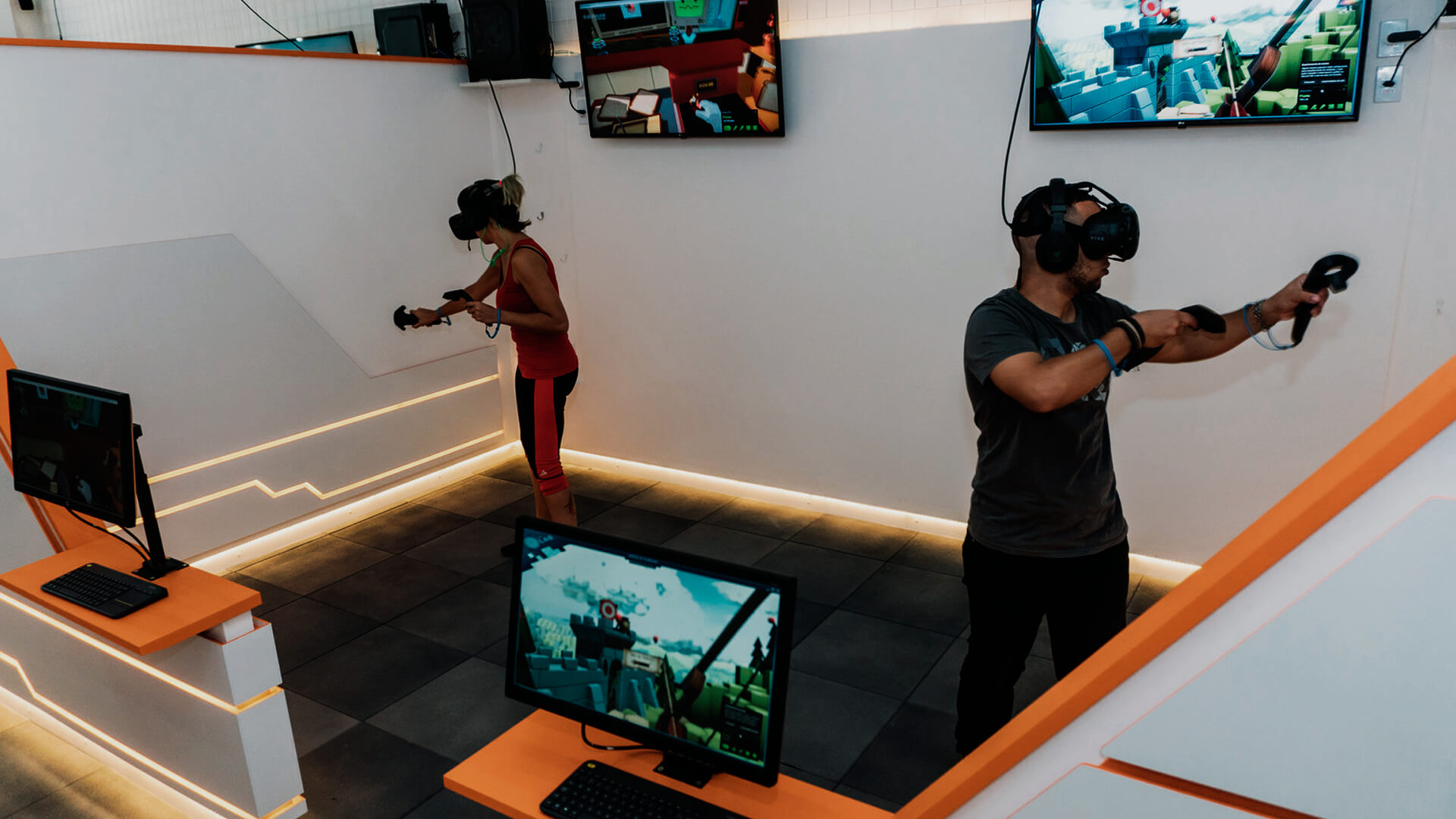 A imagem é atual e mostra um casal utilizando a realidade virtual para jogar. O ambiente é uma sala com várias telas, parecendo bastante uma exposição.