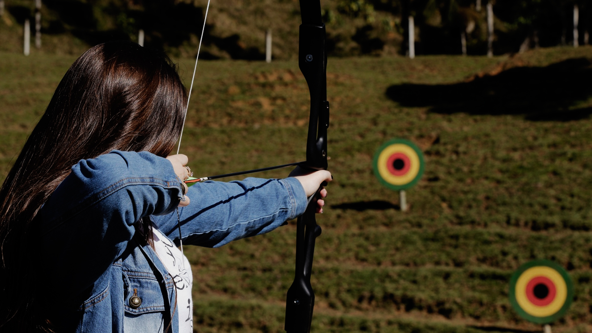 Mulher praticando arquearia. Na imagem ela está mirando o arco e flecha em um alvo. Ela está em um campo aberto e os alvos localizados em cima da grama.