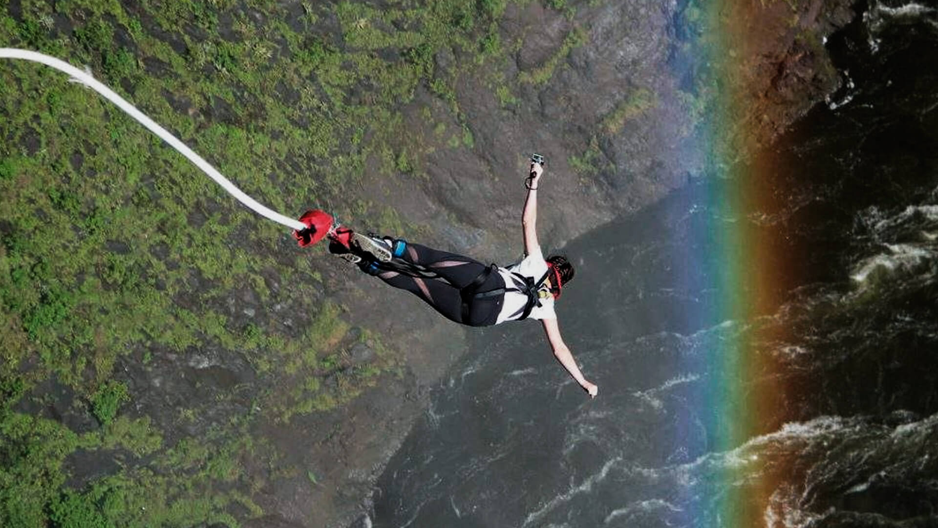 Mulher saltando de bungee jump com os braços abertos. A paisagem tem diversas árvores, um rio e um arco-íris.