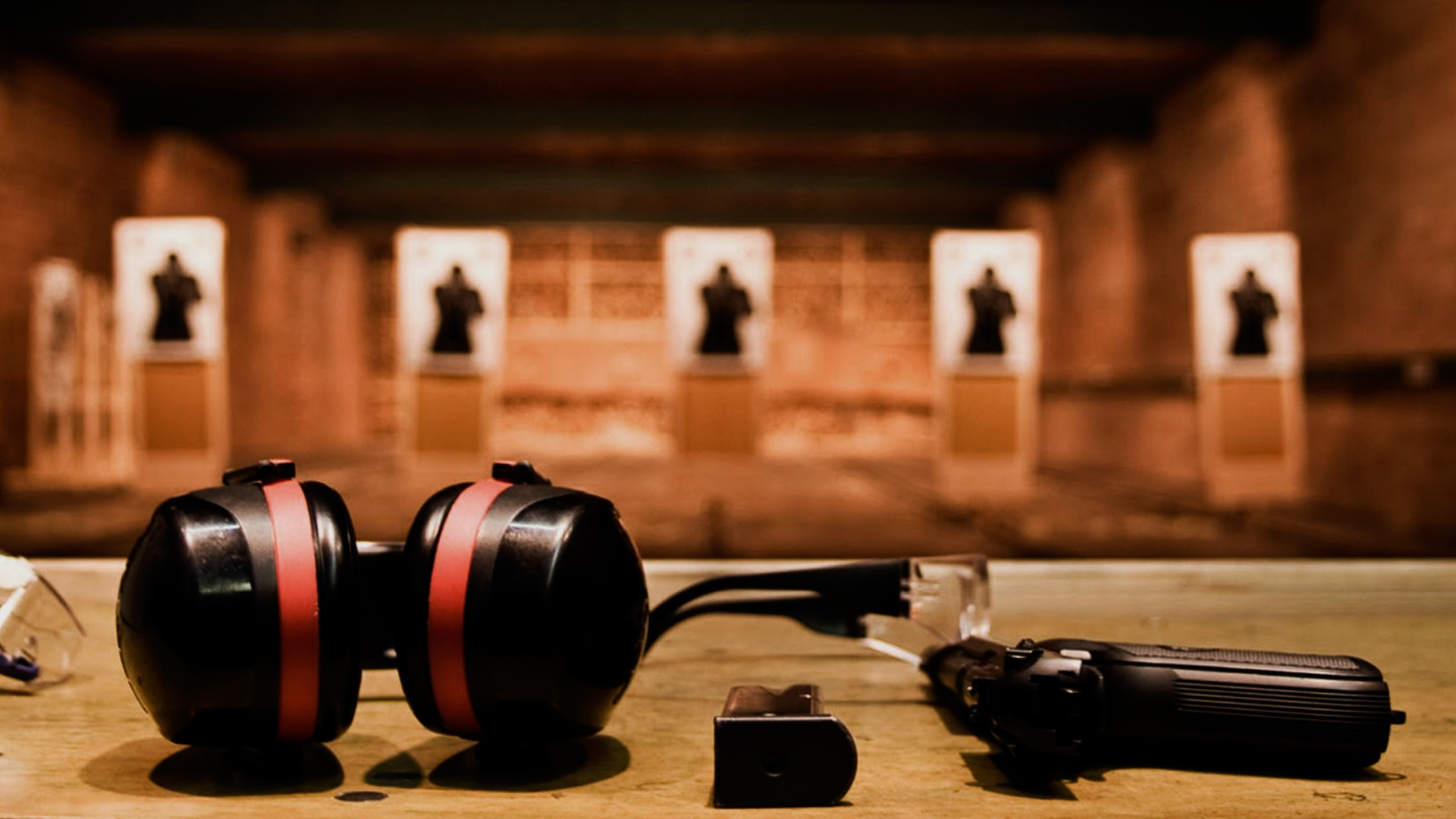 Na imagem tem uma mesa a frente, em um ambiente de estande de tiro. Sobre a mesa tem um protetor auricular, uma arma, um pente de arma e óculos de proteção. Ao fundo tem 5 alvos para realizar os disparos.