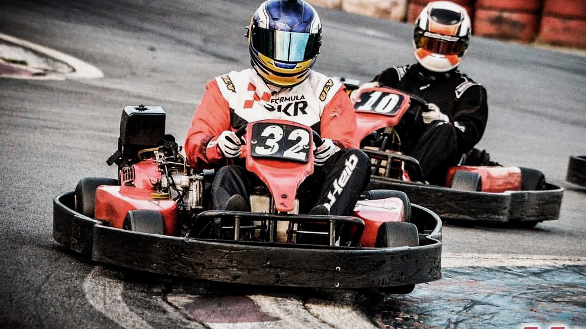 Dois pilotos fazendo uma curva na pista de kartódromo. O piloto da frente está passando em cima da zebra na curva.