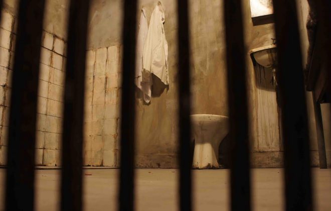 Na imagem aparecem grades e ao fundo tem uma pequena prisão, com uma pia, um vaso sanitário e uma toalha no varal.