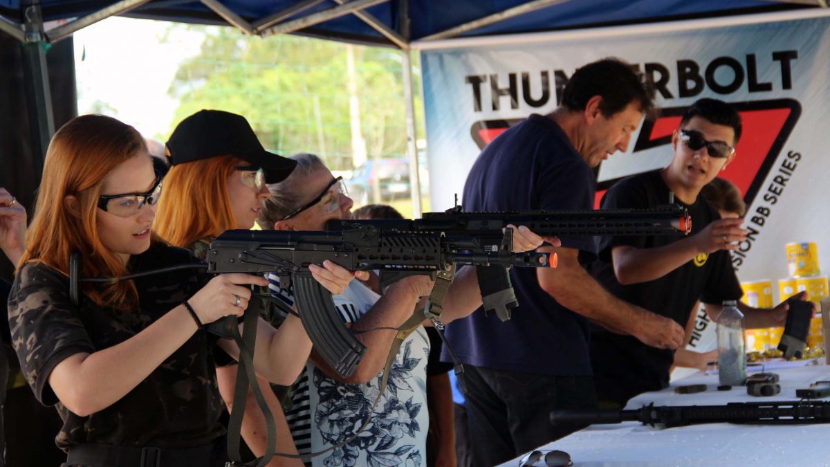 Na imagem há um estande de tiro, com três mulheres segurando armas de airsoft, prontas para atirar.