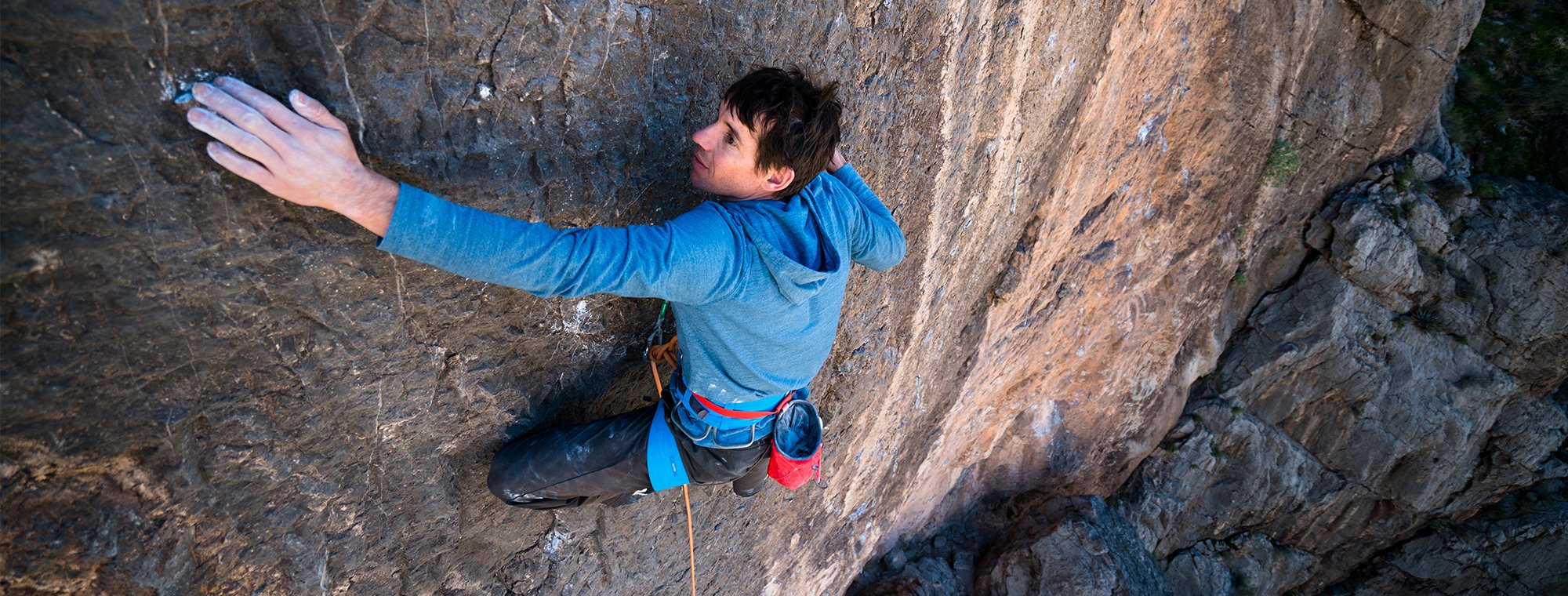 Na imagem, o alpinista Alex Honnold está escalando um paredão, ele usa um casaco azul e está utilizando corda, já que é uma escalada teste.