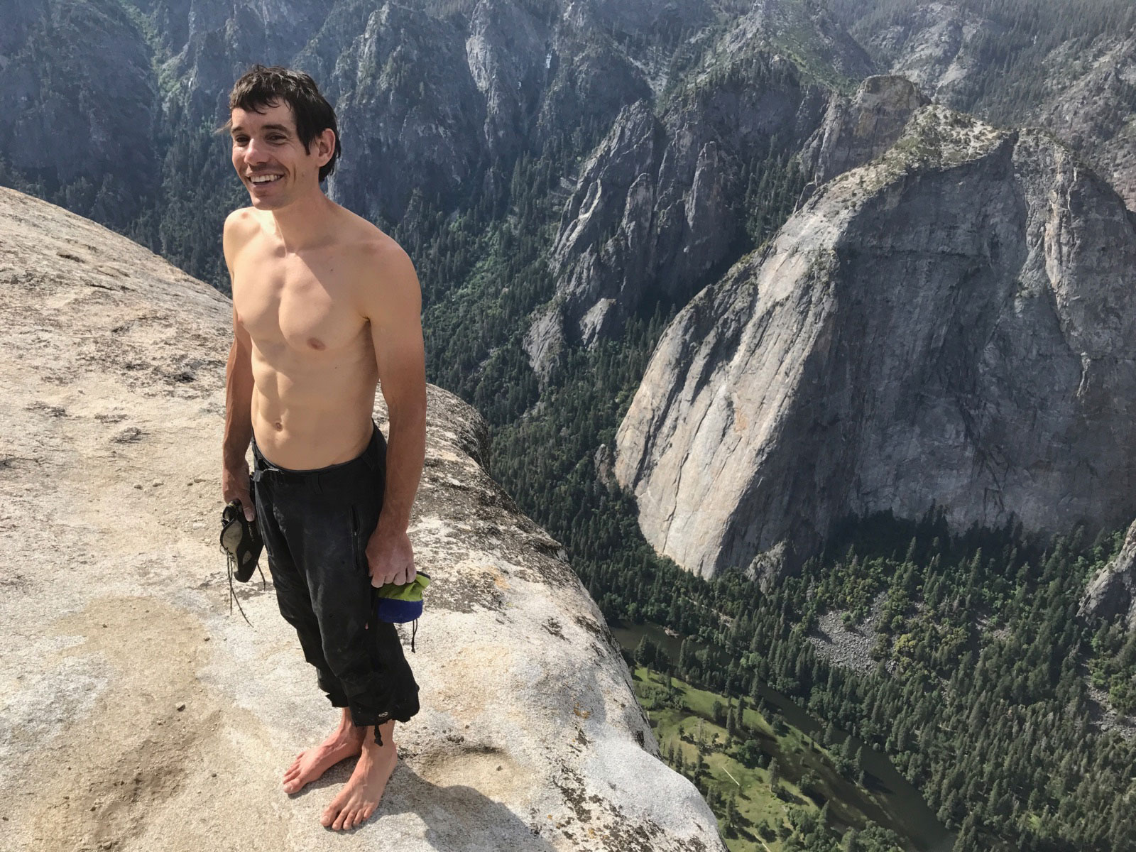 Na imagem, Alex Honnold está descalço, segurando seu tênis de escalada. Ele está no topo de um paredão, e sorri para a câmera.