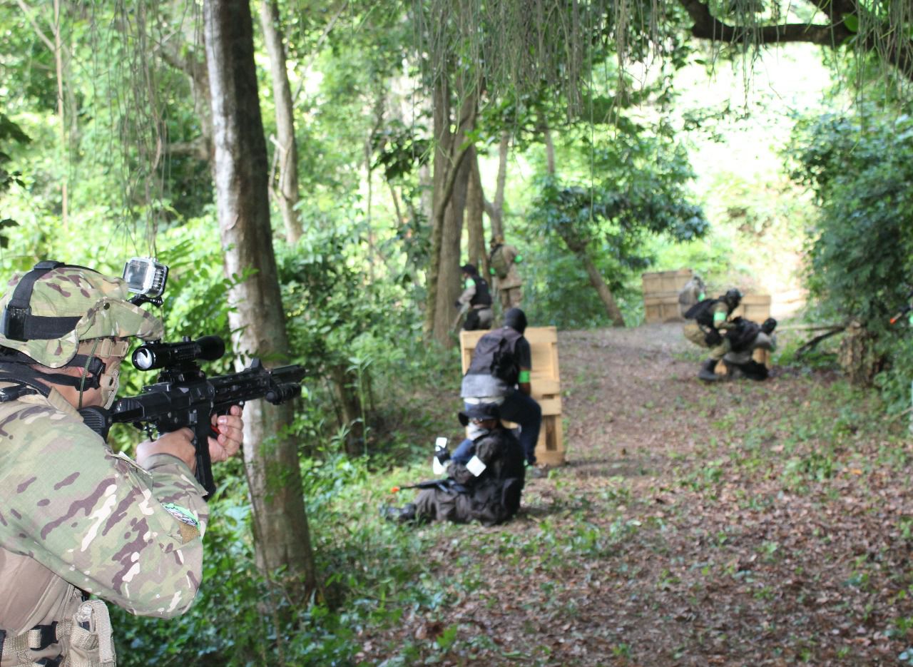 Na imagem, três jogadores de airsoft se posicionam atrás dos obstáculos e apontam suas armas.