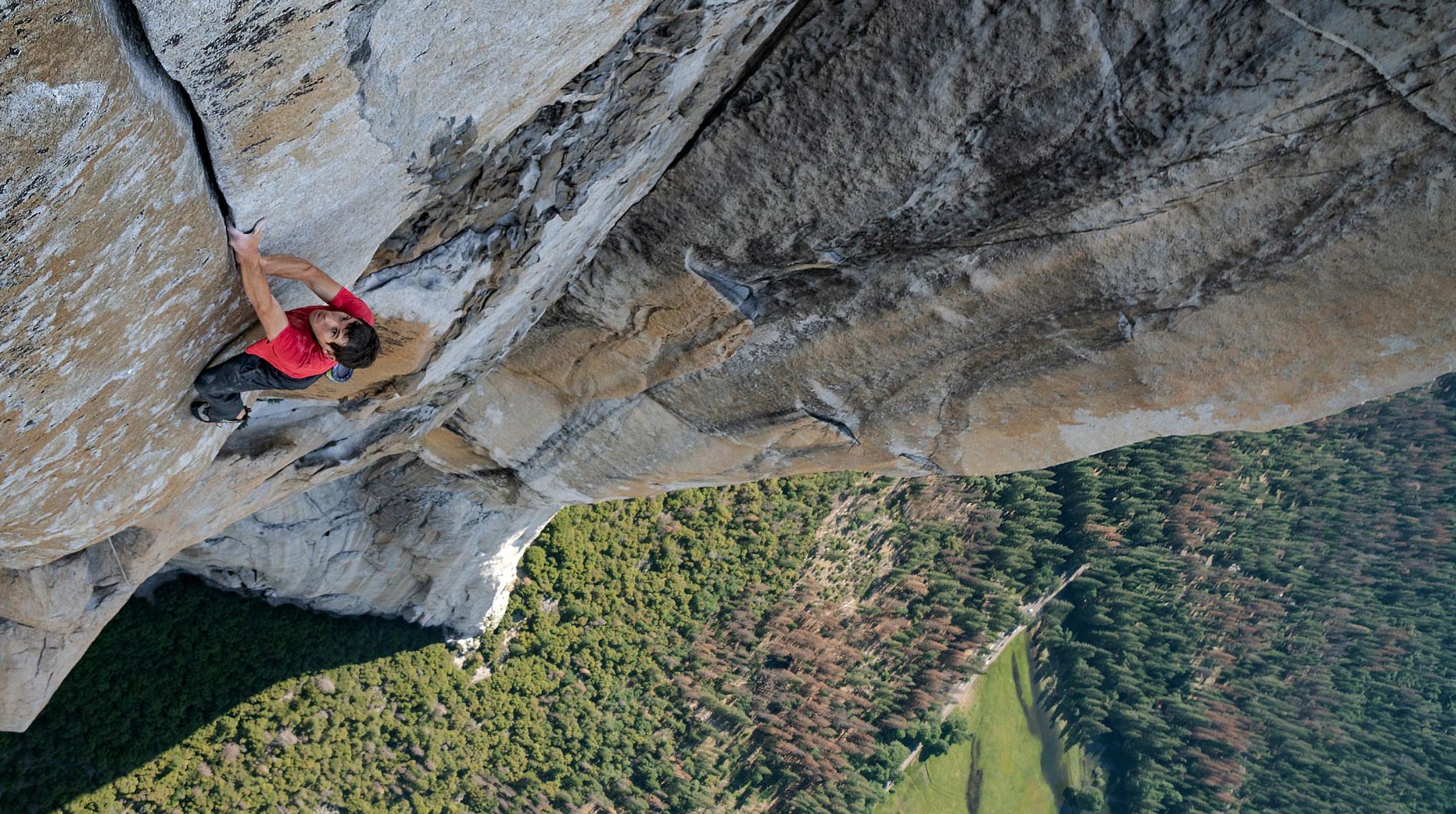 Na imagem, Alex Honnold escala um paredão gigante. Ele está com as mãos entre um pequeno vão, sem protecão alguma. 