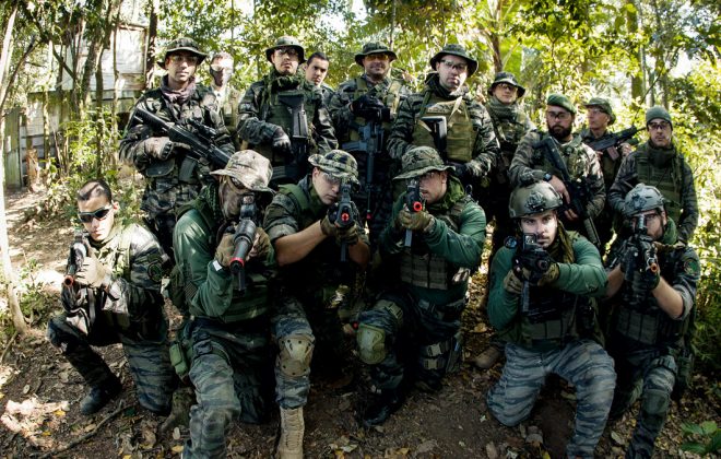 Na imagem, temos um time de airsoft composto por homens, todos trajados com roupas para a prática. Ao fundo, um campo no estilo Selva.