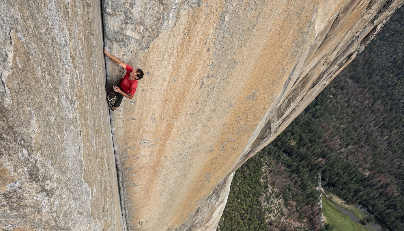 Na imagem, Alex aparece escalando um paredão enorme. O alpinista está com as mãos entre uma abertura da rocha.