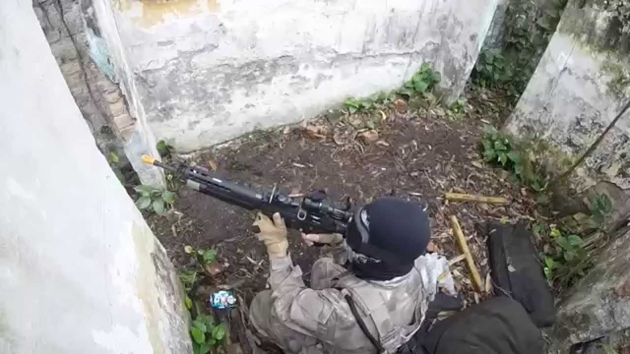 Na imagem, um homem com as vestimentas próprios para o airsoft, segura uma arma e aponta para fora do seu esconderijo.