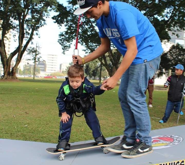 Na imagem, um garoto que está sendo segurado pelo instrutor e por cordas, anda de skate adaptado.