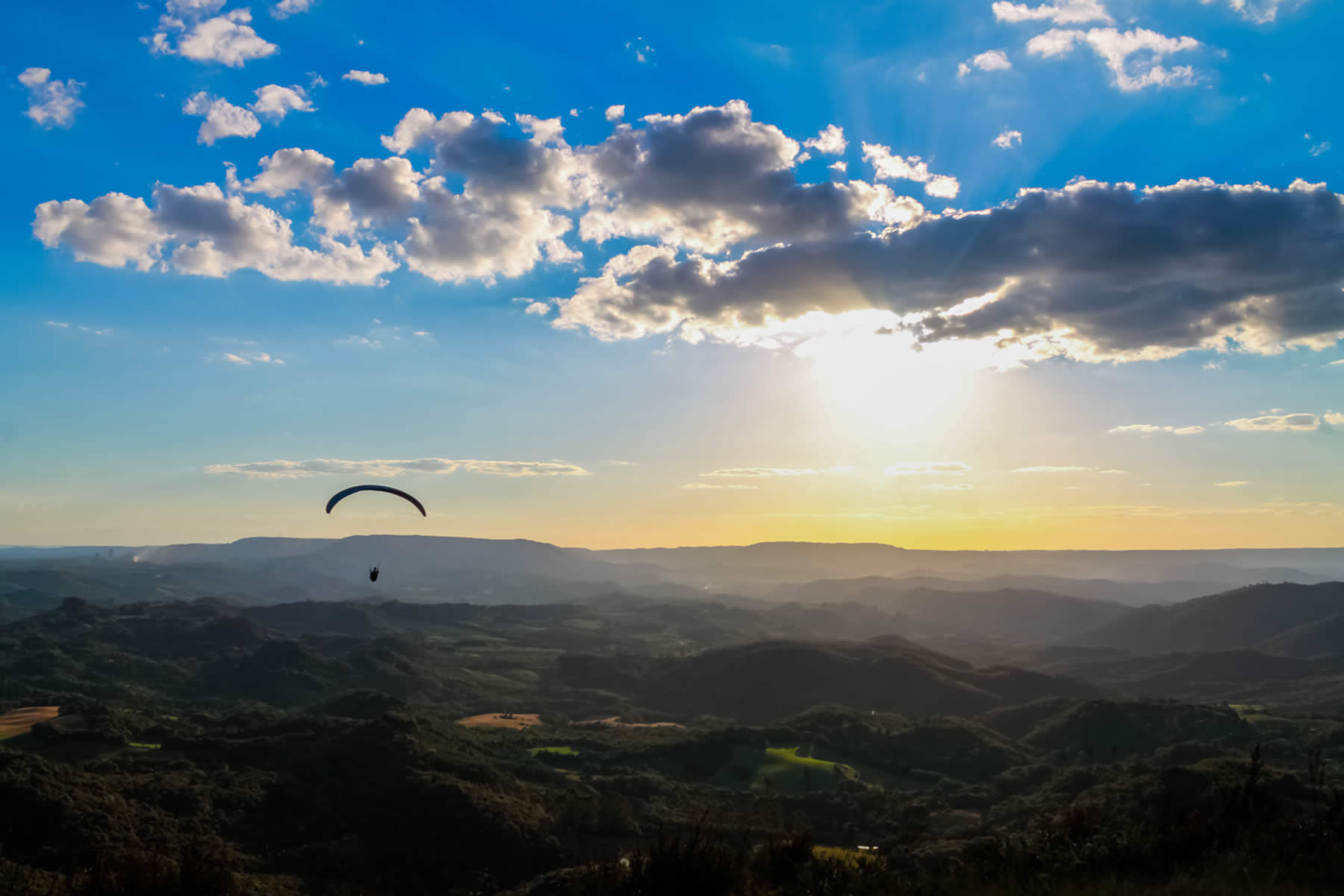 Na imagem, temos a vista do Morro do Cal. O céu está muito azul e tem alguém de parapente voando sob os morros menores.