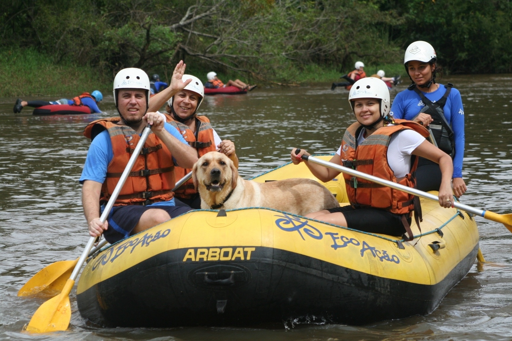 Na imagem, um grupo de 4 pessoas e 1 cachorro, estão sobre um bote em uma corredeira de água.