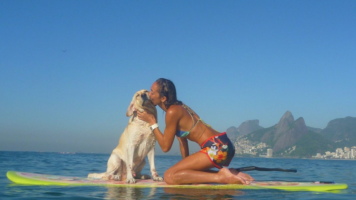 Na imagem, uma moça e seu cãozinho estão sobre as águas, em cima de uma prancha de stand up paddle. Na foto a moça está dando um beijinho no seu cachorro.