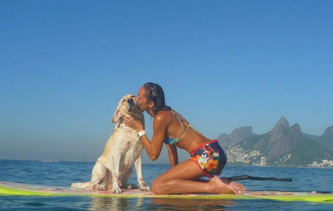 Na imagem, uma moça e seu cãozinho estão sobre as águas, em cima de uma prancha de stand up paddle. Na foto a moça está dando um beijinho no seu cachorro.