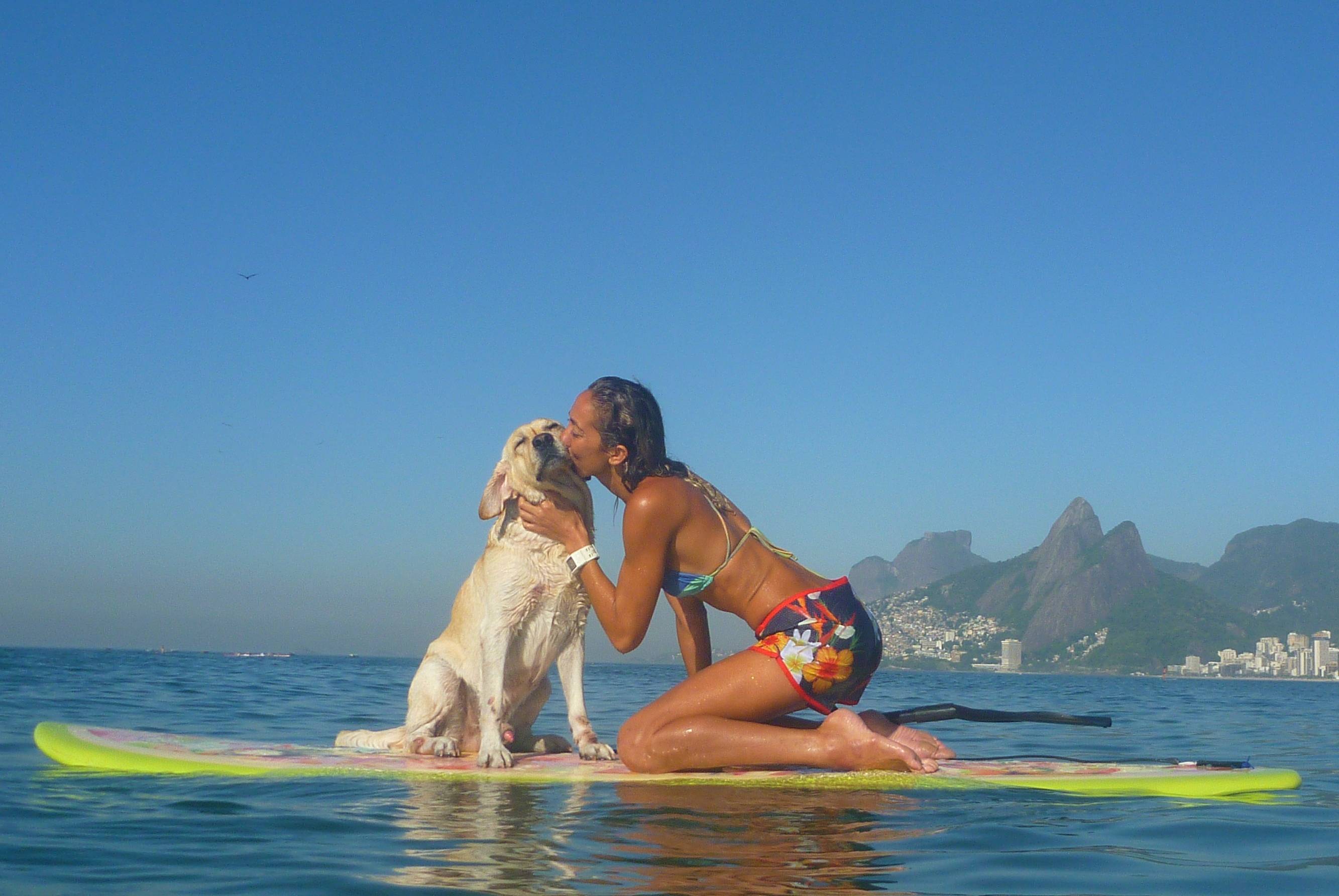 Na imagem, uma moça e seu cãozinho estão sobre as águas, em cima de uma prancha de stand up paddle. Na foto a moça está dando um beijinho no seu cachorro.