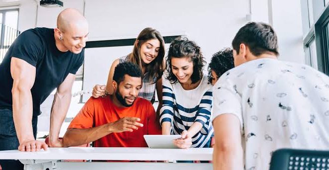 Na imagem, pessoas jovens, dois homens e duas mulheres se reunem em uma mesa de trabalho.