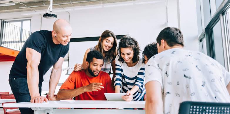 Na imagem, pessoas jovens, dois homens e duas mulheres se reunem em uma mesa de trabalho.