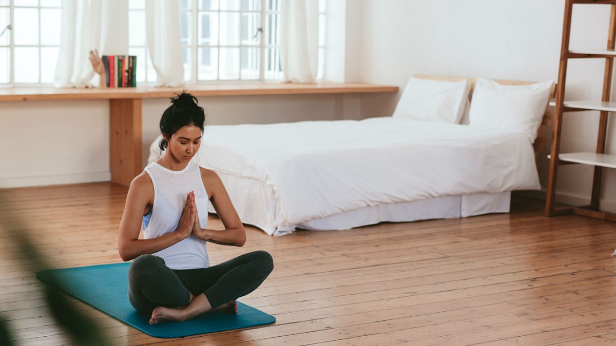 Na imagem, uma moça faz a posição clássica da meditação. Sentada no chão, em cima de um tapete de yoga, ela cruze as pernas e junta as mãos.