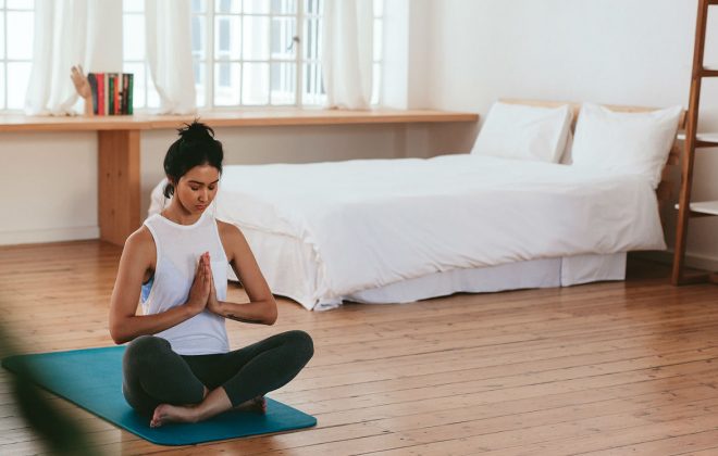 Na imagem, uma moça faz a posição clássica da meditação. Sentada no chão, em cima de um tapete de yoga, ela cruze as pernas e junta as mãos.