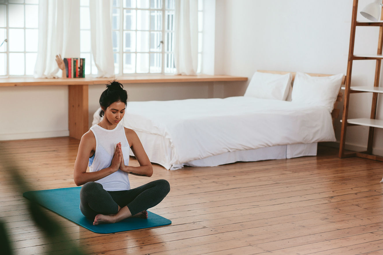 Na imagem, uma moça faz a posição clássica da meditação. Sentada no chão, em cima de um tapete de yoga, ela cruze as pernas e junta as mãos.