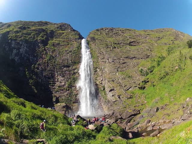 Na imagem, há uma cachoeira em meio dois montes. Ela forma uma piscina natural onde as pessoas se banham.