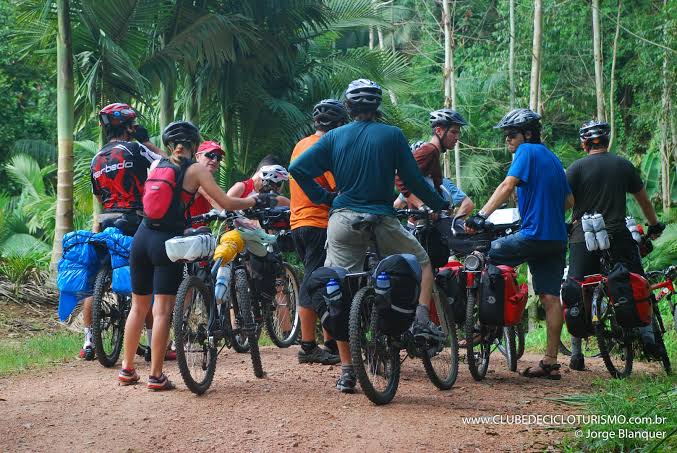 Na imagem, um grupo de ciclistas se reúnem em uma trilha em meio a natureza.