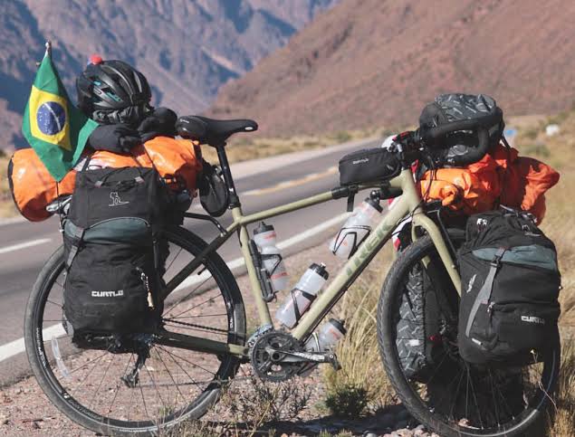 Na imagem, uma bicicleta com uma pequena bandeira do Brasil pendurada, é fotografada junto de algumas mochilas por perto.