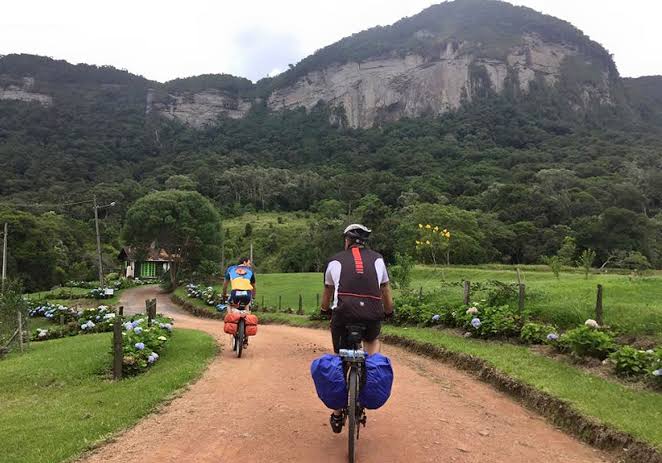 Na imagem, dois ciclistas andam por uma estrada de terra em meio a natureza.