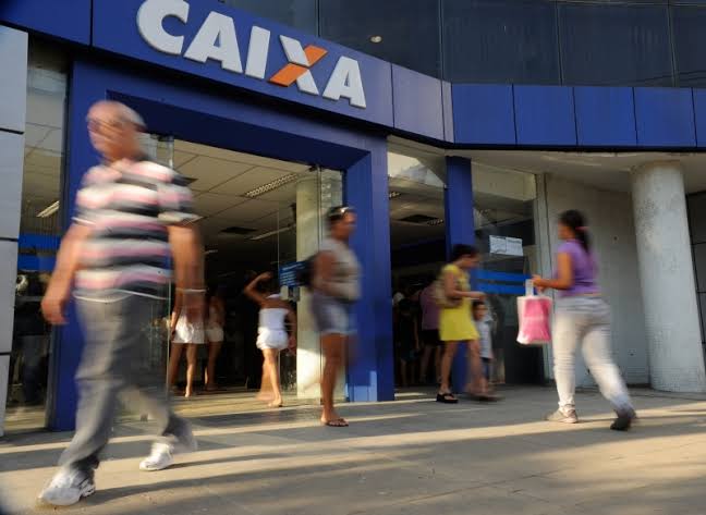 Na imagem, há um estabelecimento com uma porta grande com o leiteiro "CAIXA” nome do banco a qual ela pertence. Enquanto isso, várias pessoas andam em frente a ela.