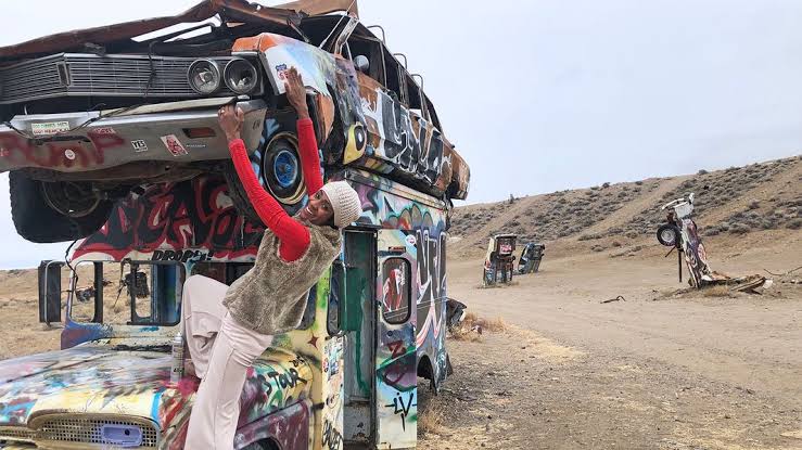 Na imagem há um deserto e carros grafitados e depredados. Em foco, uma moça posa para a foto em um desses carros.